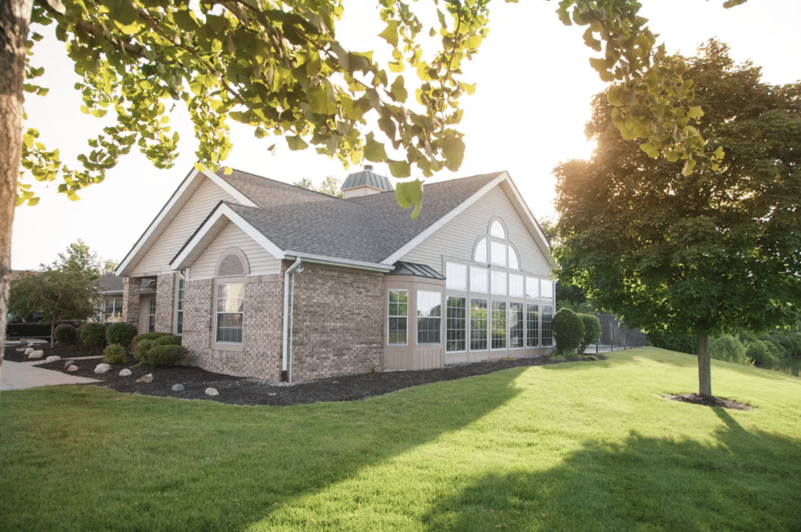 Single-story brick house with large windows, surrounded by a well-maintained lawn and trees, photographed in bright sunlight.
