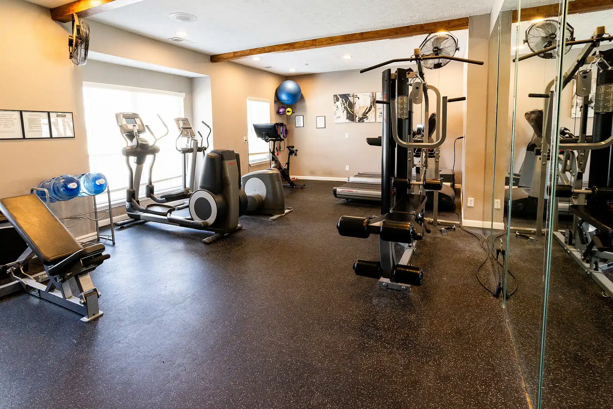 A fitness room with treadmills, ellipticals, weight machines, an exercise bench, mirrors, and wall-mounted fans.